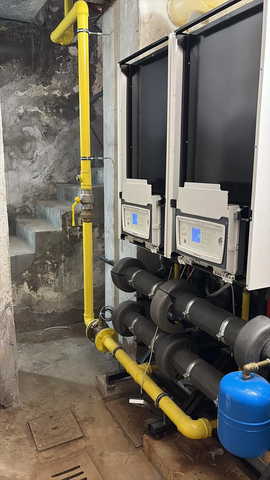 Industrial pipes and control panels with yellow gas piping and a blue pressure tank in a basement with concrete stairs.