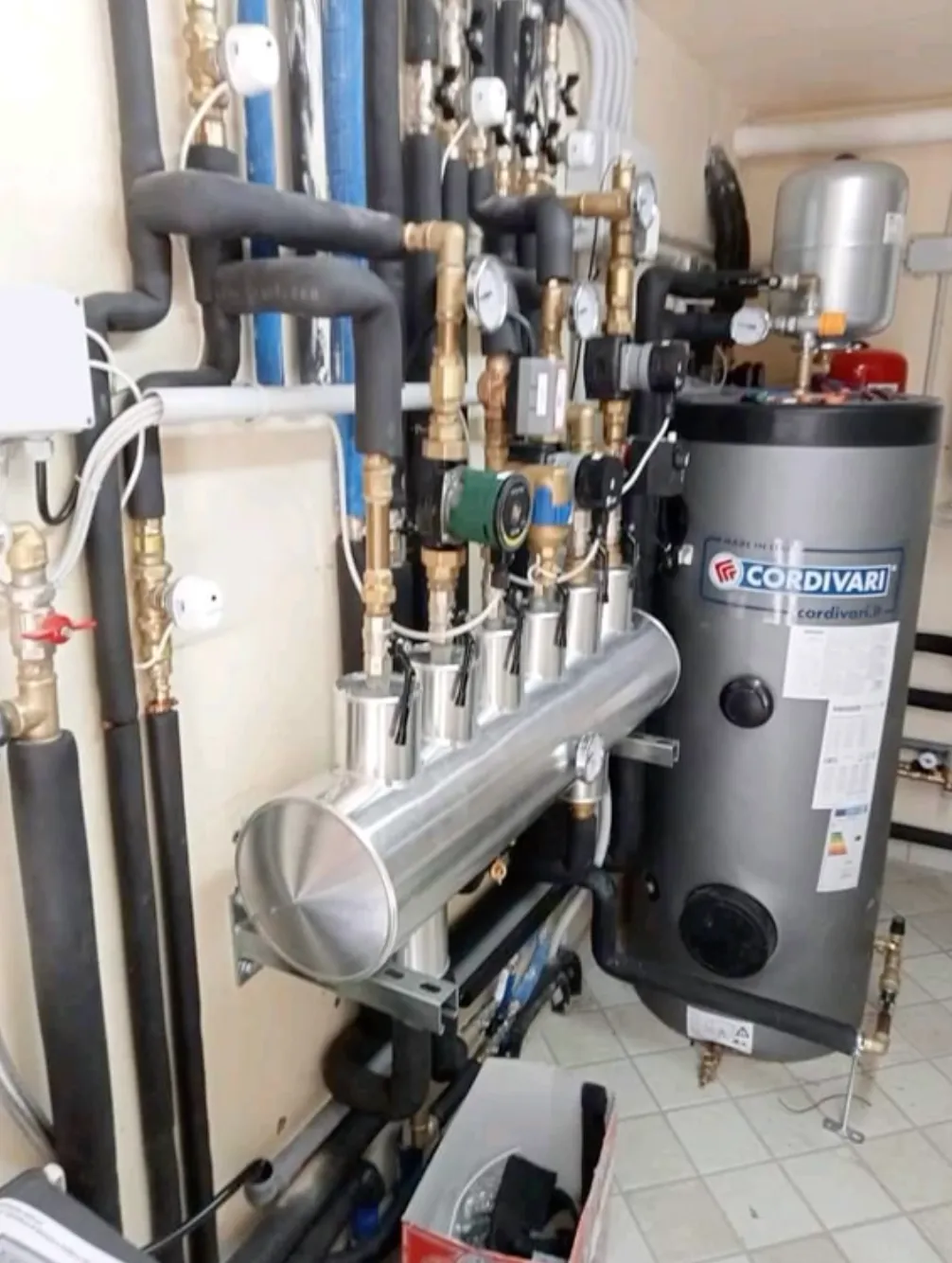 Industrial heating system with a Cordivari water heater, multiple insulated pipes, gauges, and brass fittings installed in a tiled utility room.