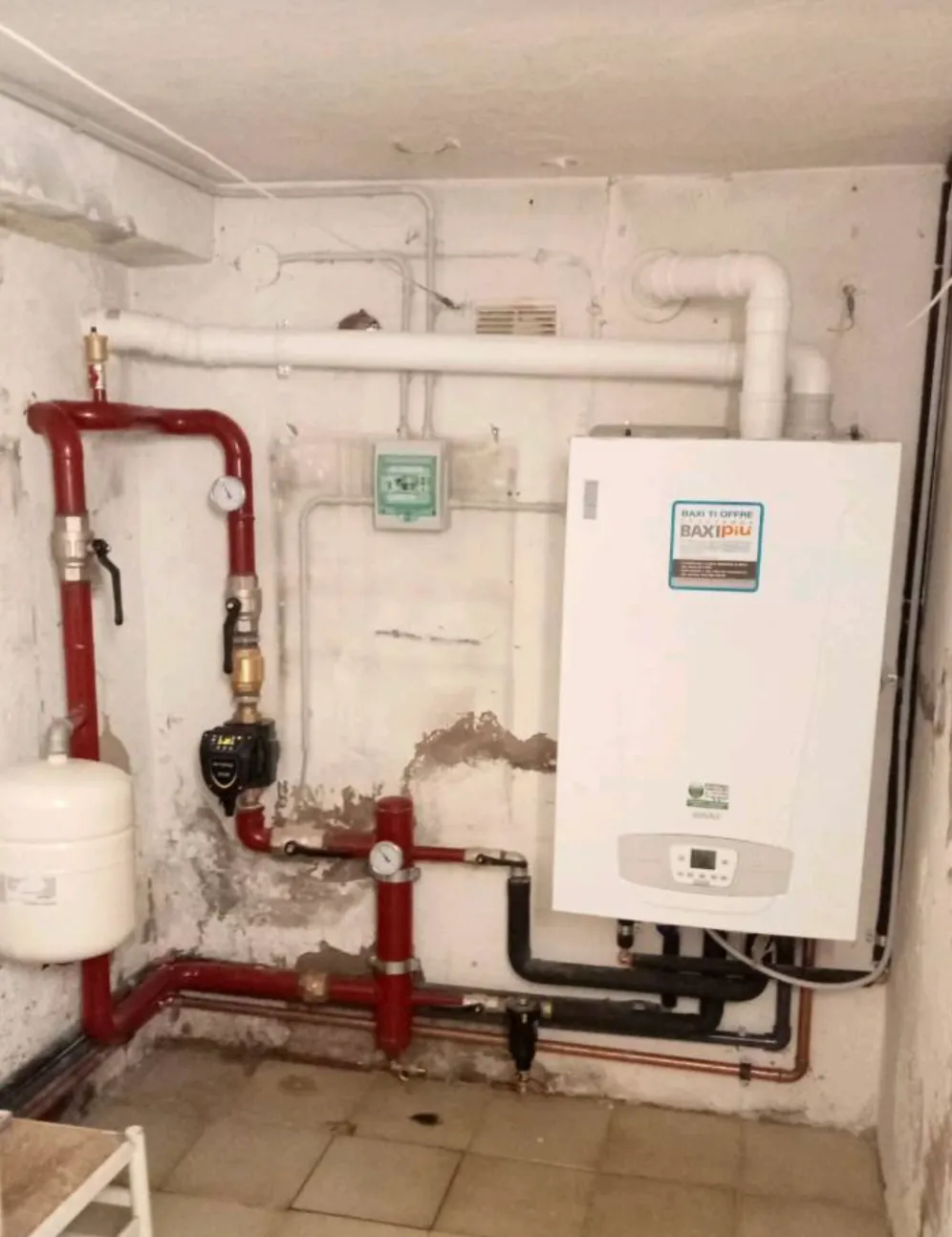 Wall-mounted boiler system with connected red and black pipes, pressure gauges, and a white expansion tank in a basement room.