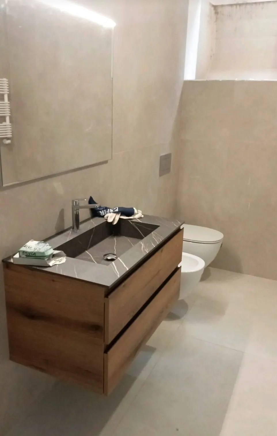 Modern bathroom with beige tiles, wall-mounted wooden vanity with a gray marble sink, a mirror above, and a white toilet and bidet.