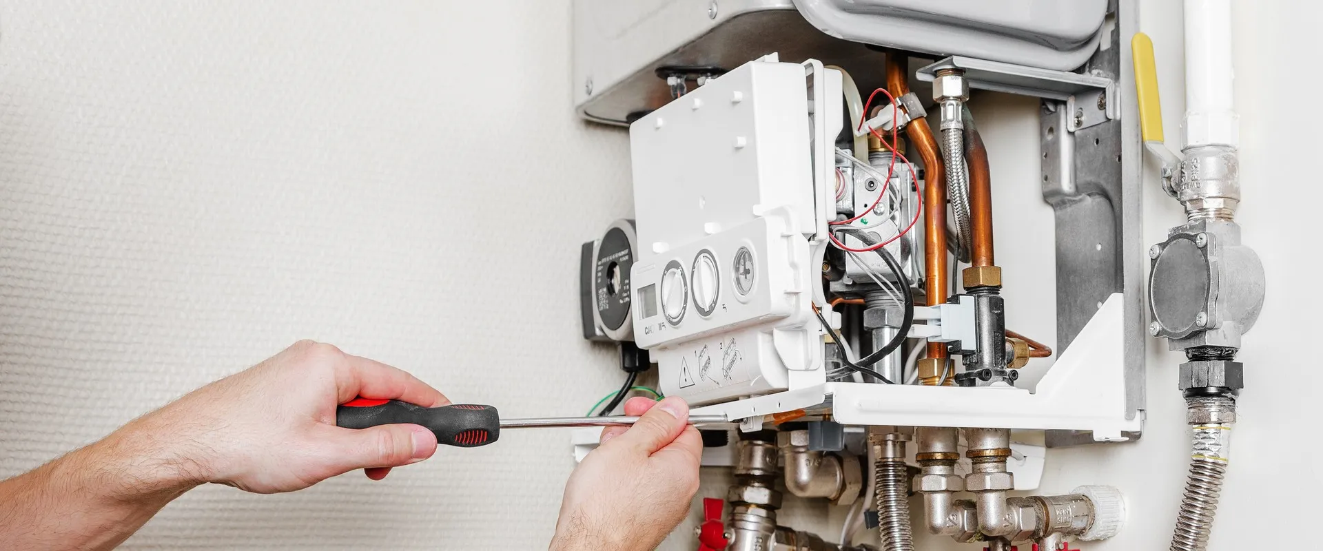 Hands using a screwdriver to repair or adjust a wall-mounted boiler with exposed pipes and controls.